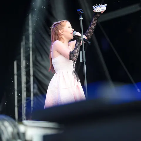 A West End Kids performer singing on an outdoor stage, holding a crown aloft
