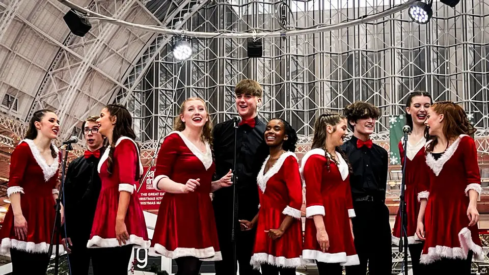 The West End Kids troupe singing in festive costumes on a stage under a glass roof