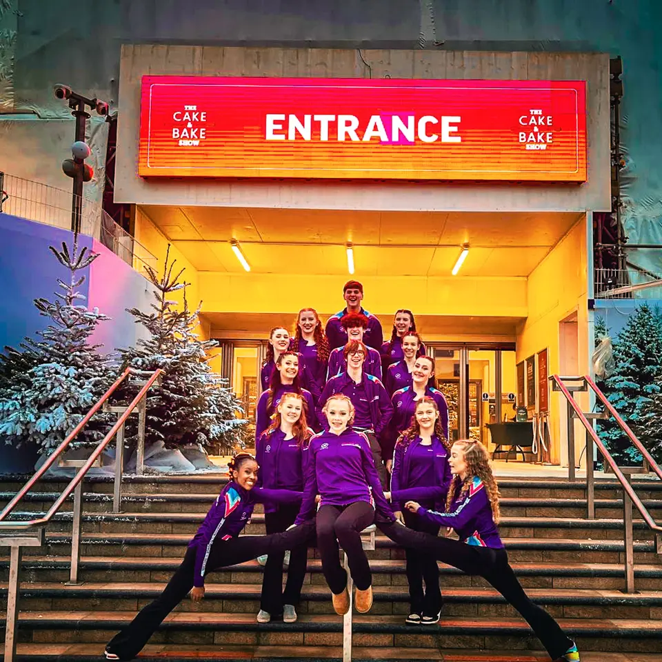 The West End Kids troupe in purple team jackets on the steps beneath the Cake and Bake Show entrance sign