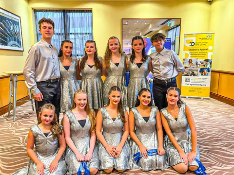 Eleven West End Kids performers in silver sequin dresses and satin shirts, posed in two rows beside a Dudley Group NHS Charity Children's Appeal banner